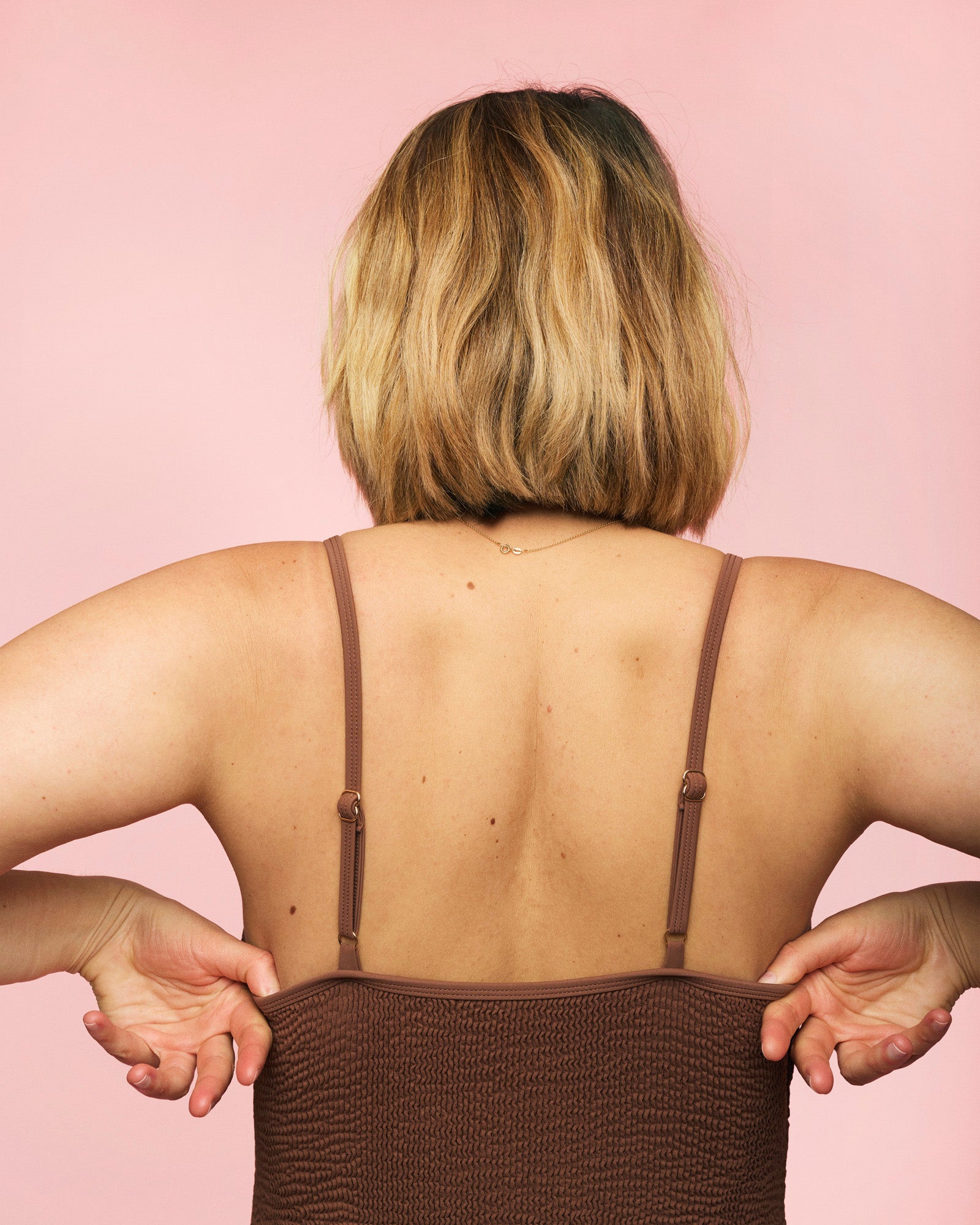 Kvinde står med rykken til iført momkind gravid badedragt i farven chocolate /brun. Man kan se stropperne bagfra.