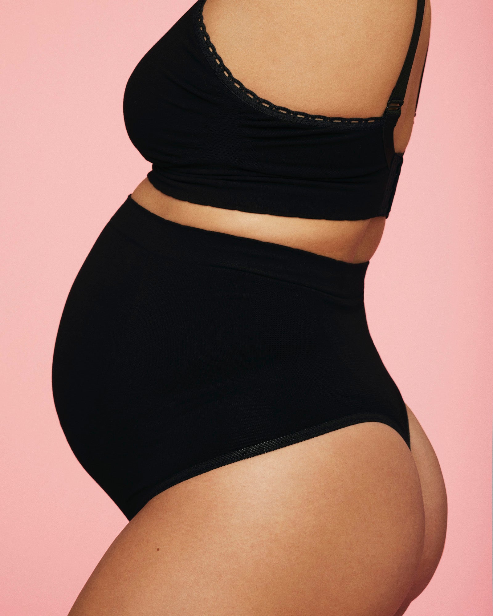 Kvinde der er iført momkinds belly support gravid g-streng og amme-bh i farven sort. Hun står med siden til og billedet er skåret, så man kun ser hendes torso.