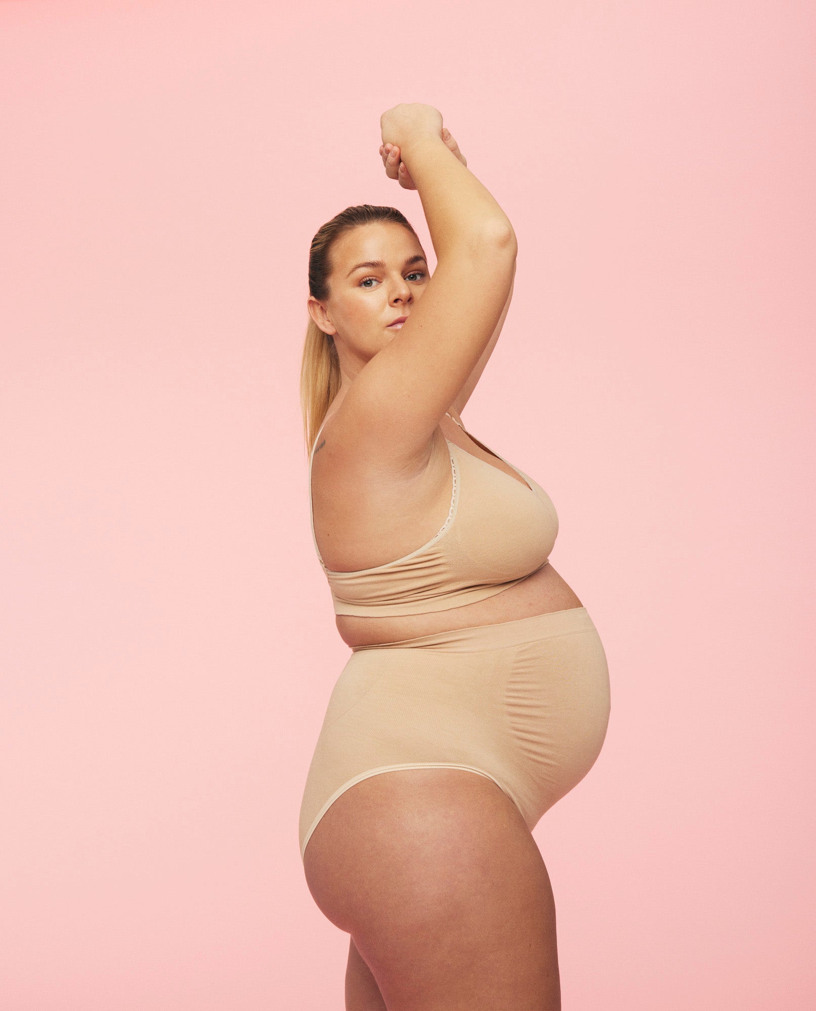 Gravid kvinde iført momkinds belly support gravidtrusser og ammebh i farven sand. Hun står med siden til og løfter armene op over hovedet.