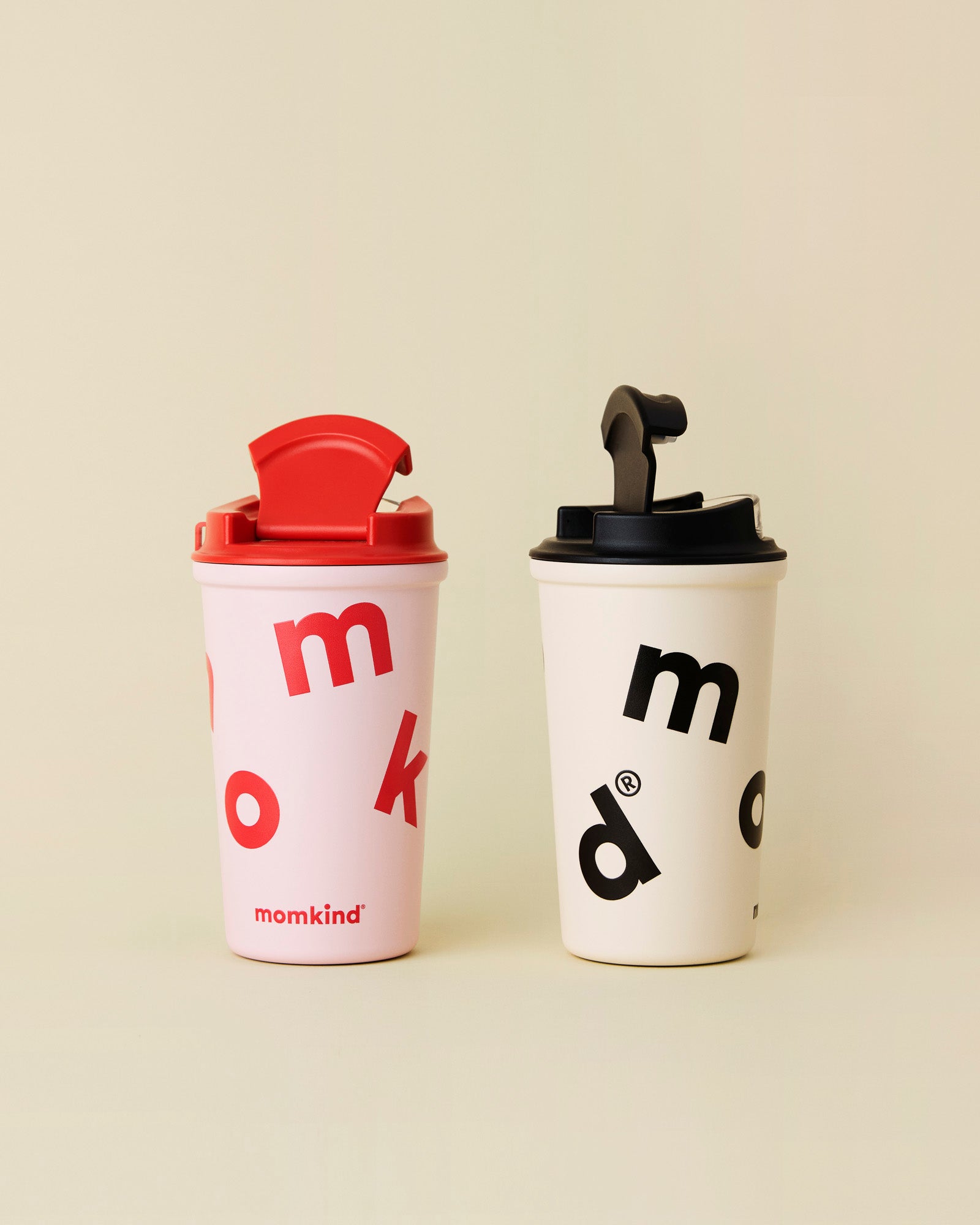 To to go termokrus med låg fra momkind. Den ene er cotton candy / lyserød med rød tekst, den anden er oatmeal med sort tekst. Låget er åbent på begge termokrus.