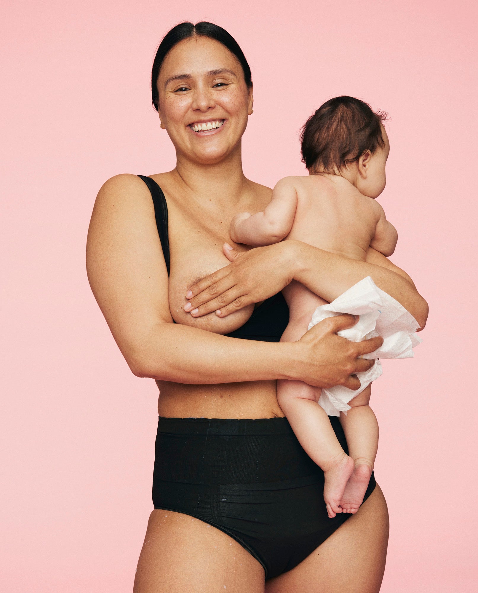 smilende kvinde har baby på den ene arm mens hun dækker sit bryst med den anden hånd og er iført sort kejsersnittrusse fra momkind