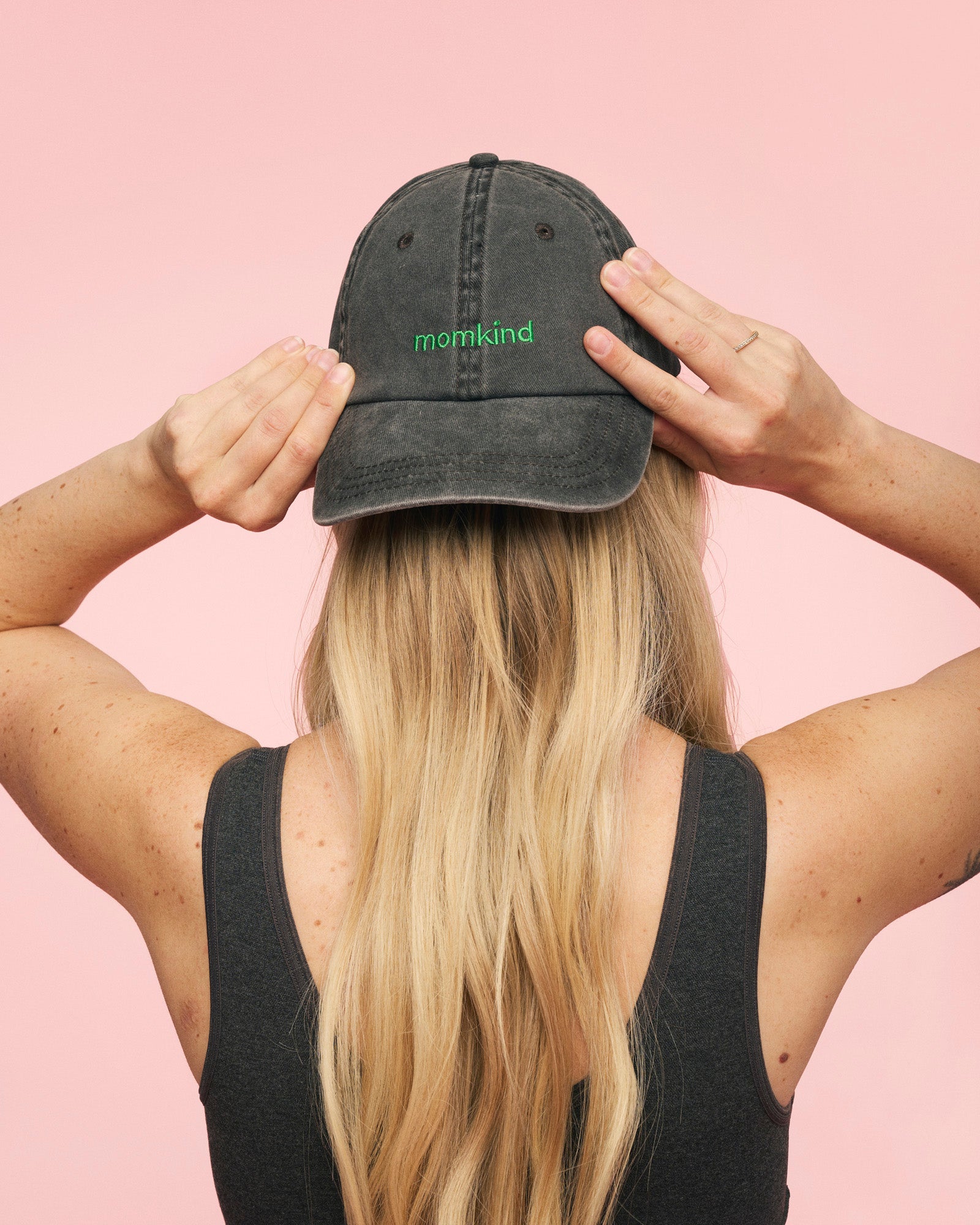 Momkind cap I sort med grønt logo. Den sidder omvendt på hovedet af en kvinde som holder begge hænder på.