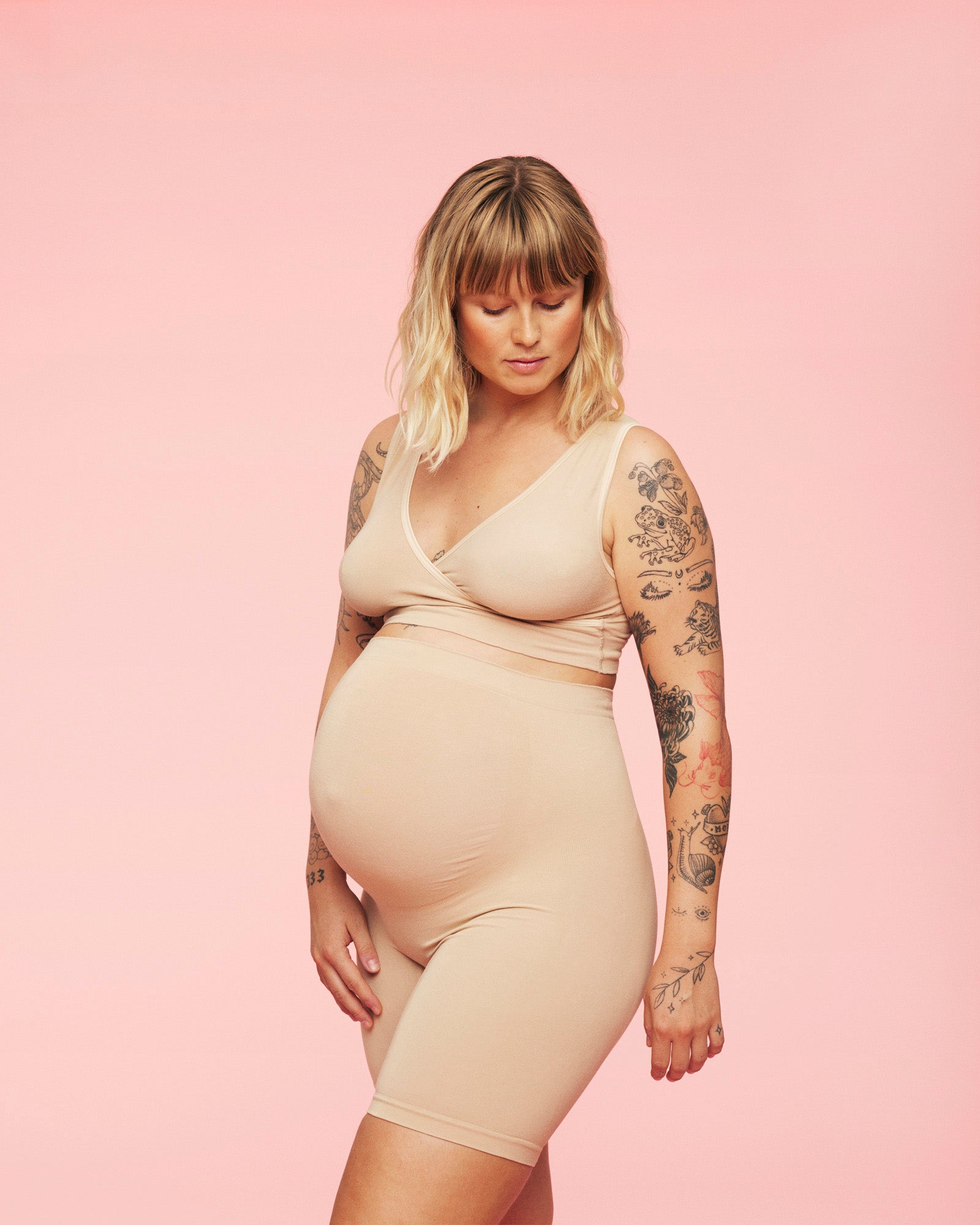 stående gravid kvinde kigger ned på maven iført momkind venteshorts til gravide og crossover amme bh i farven sand